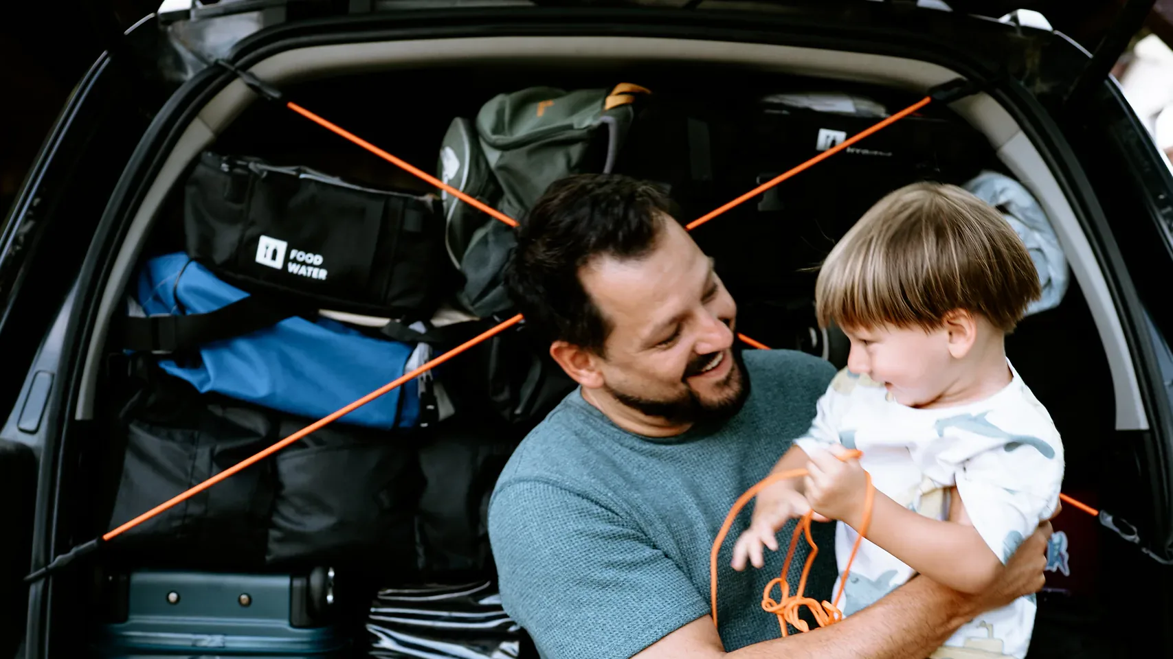 Tata z synkiem w otwartym bagażniku, bagaż za nimi spięty na krzyż linkami elastycznymi HELP BAG