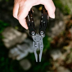 Dłoń trzyma multitool HELP BAG z rozłożonymi kombinerkami skierowanymi w dół, w tle ogród