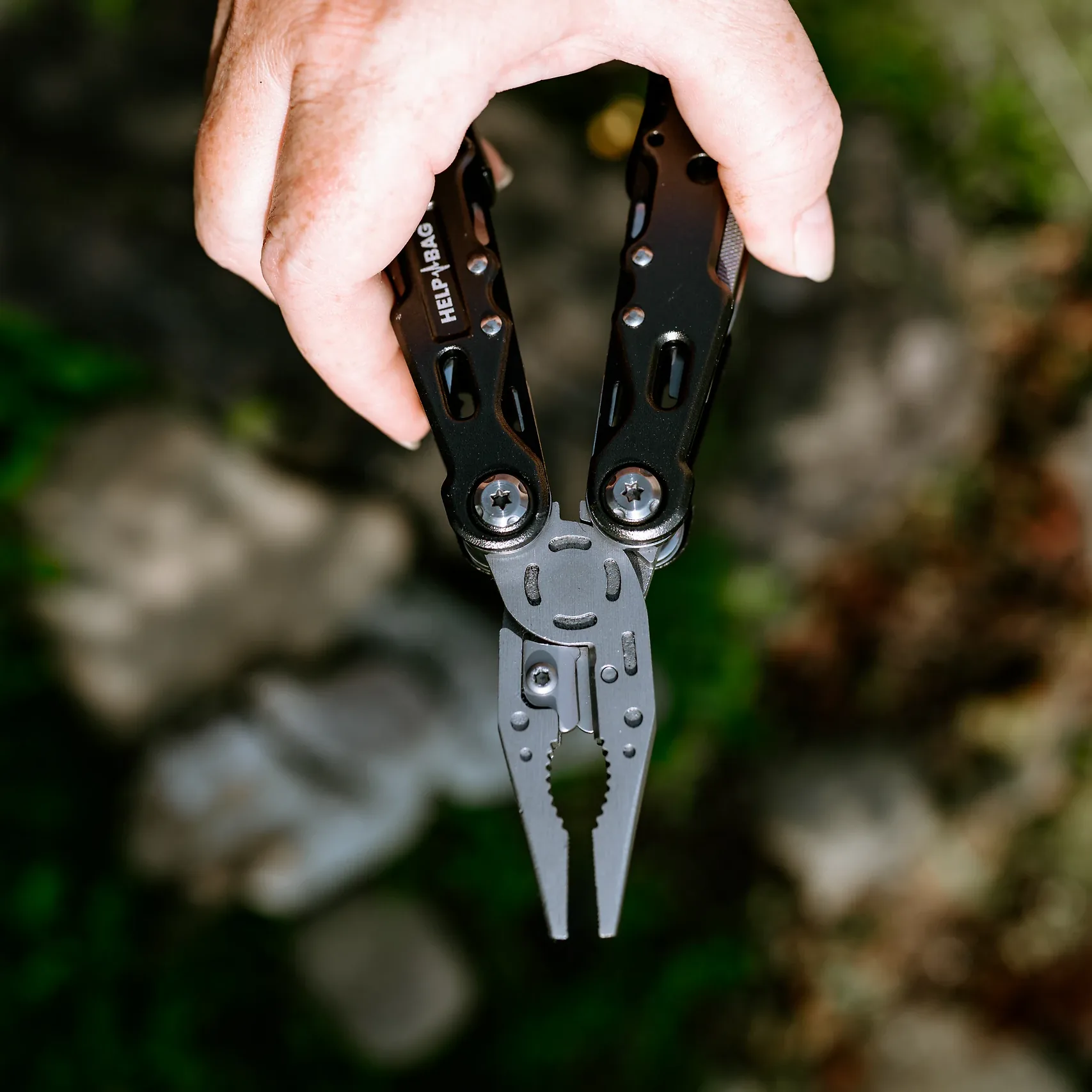 Dłoń trzyma multitool HELP BAG z rozłożonymi kombinerkami skierowanymi w dół, w tle ogród