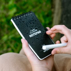 Dłonie dziecka z ołówkiem na mokrej okładce notesu wodoodpornego HELP BAG ALL-WEATHER NOTEPAD