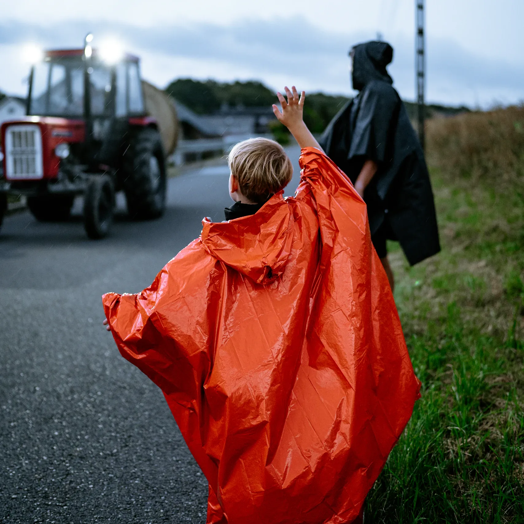 Chłopiec w poncho awaryjnym HELP BAG macha na poboczu do traktora, pomarańczowy kolor dobrze widać