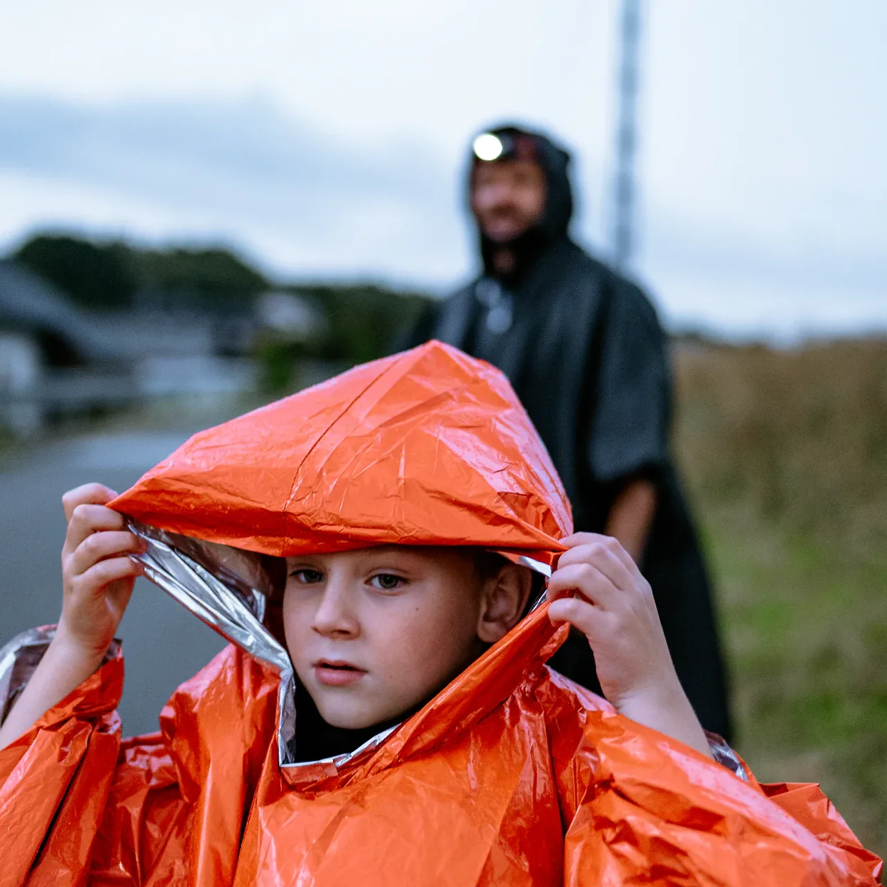Chłopiec sam naciąga kaptur pomarańczowego poncho awaryjnego HELP BAG, w tle tata w czarnym poncho