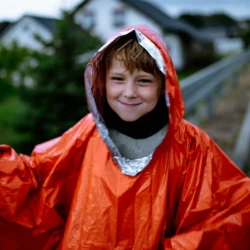 Uśmiechnięty chłopiec w kapturze poncho awaryjnego HELP BAG, widać srebrną wewnętrzną stronę folii