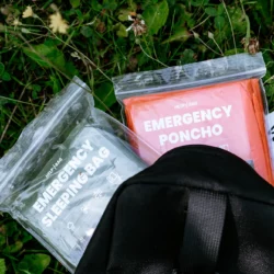 Poncho awaryjne HELP BAG w pomarańczowym opakowaniu EMERGENCY PONCHO obok śpiwora awaryjnego