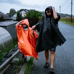 Chłopiec w pomarańczowym poncho awaryjnym HELP BAG idzie z tatą za rękę mokrą drogą w deszczu