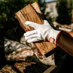 Dłoń w rękawicy skórzanej HELP BAG z owczej skóry trzyma polano przy stosie drewna