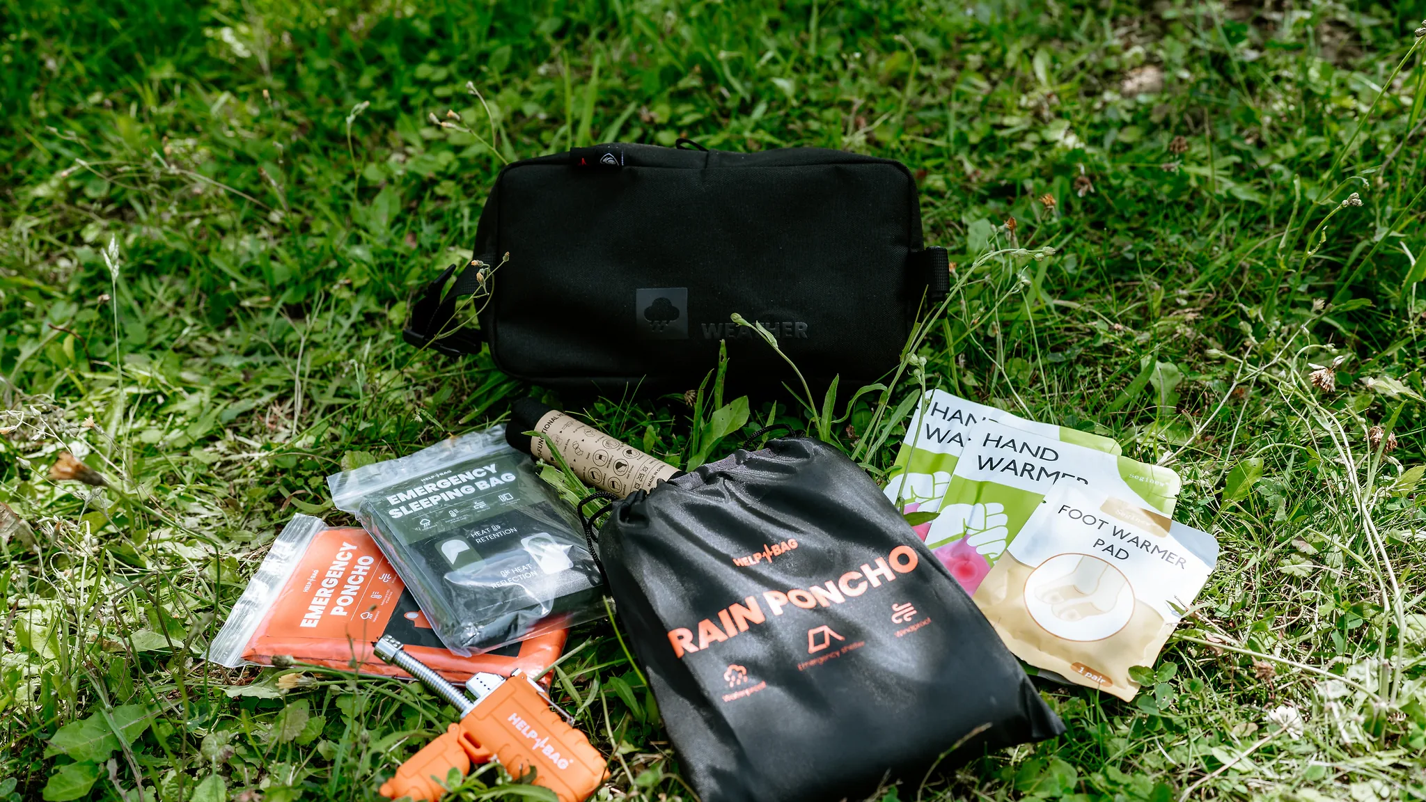 Moduł WEATHER HELP BAG na trawie, przed nim zapalniczka plazmowa, śpiwór awaryjny i ogrzewacze