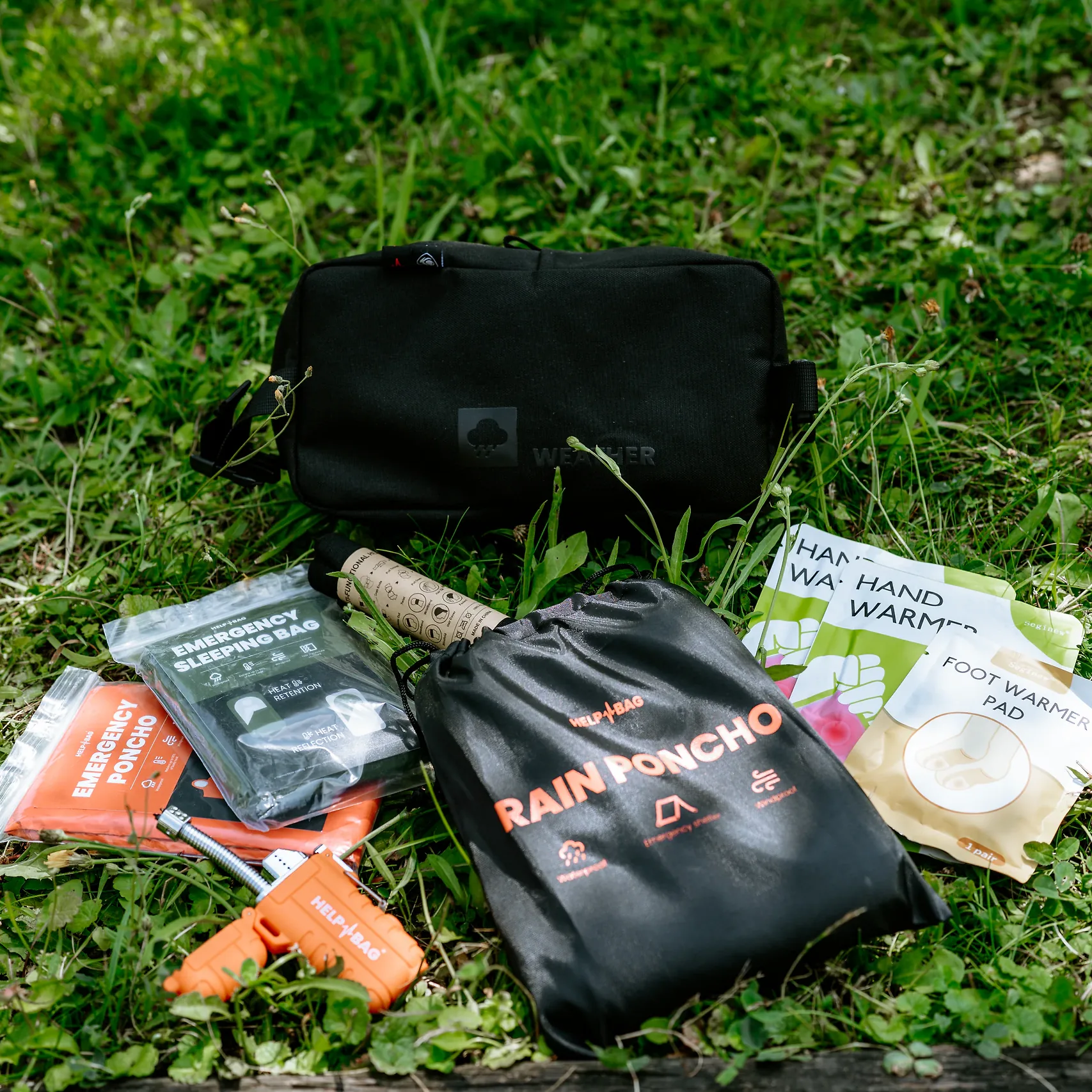 Moduł WEATHER HELP BAG na trawie, przed nim zapalniczka plazmowa, śpiwór awaryjny i ogrzewacze